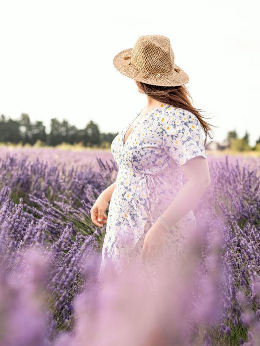 Frau im Sommerkleid von hinten, der Wind bewegt Hut und Haare über dem Lavendelfeld