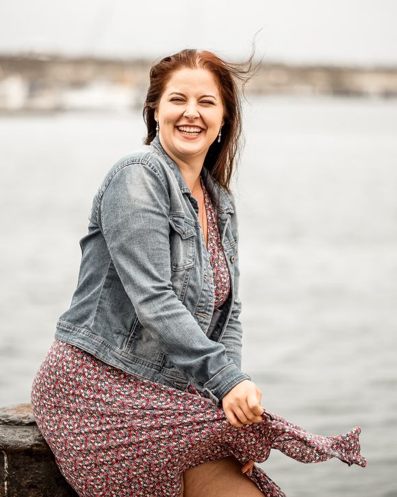 Frau in Jeansjacke sitzt lachend auf einer Mauer am Wasser