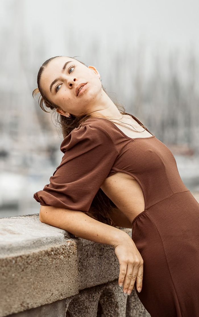 Frau im braunen Kleid lehnt an einer Mauer, im Hintergrund ein Segelhafen