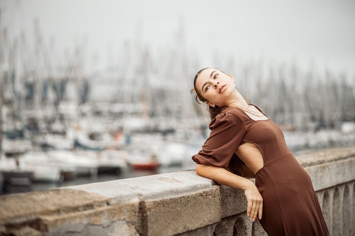 Frau im braunen Kleid lehnt an einer Mauer, im Hintergrund ein Segelhafen