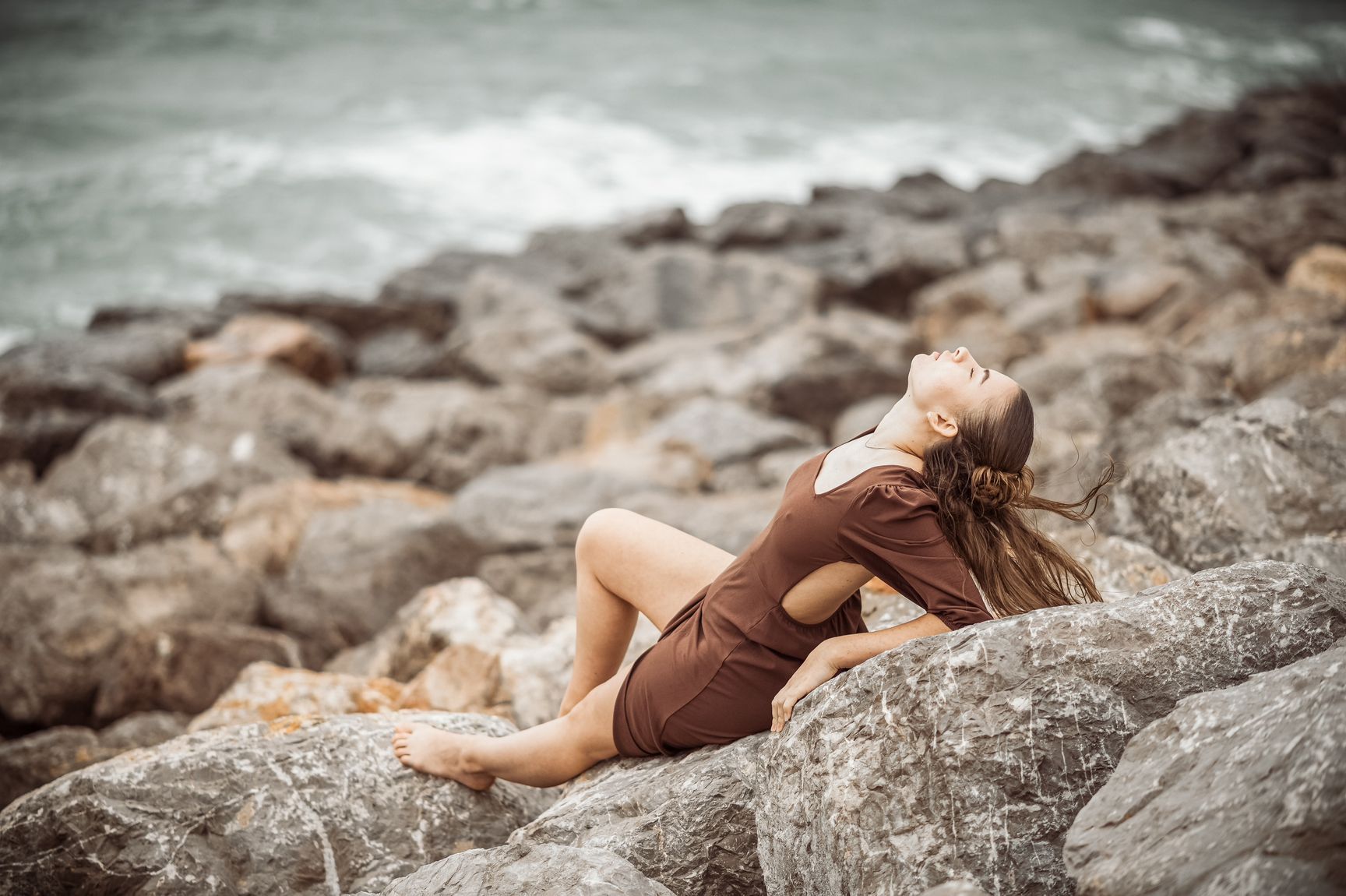 Frau im braunen Kleid liegt auf Felsen am Meer und blickt nach oben