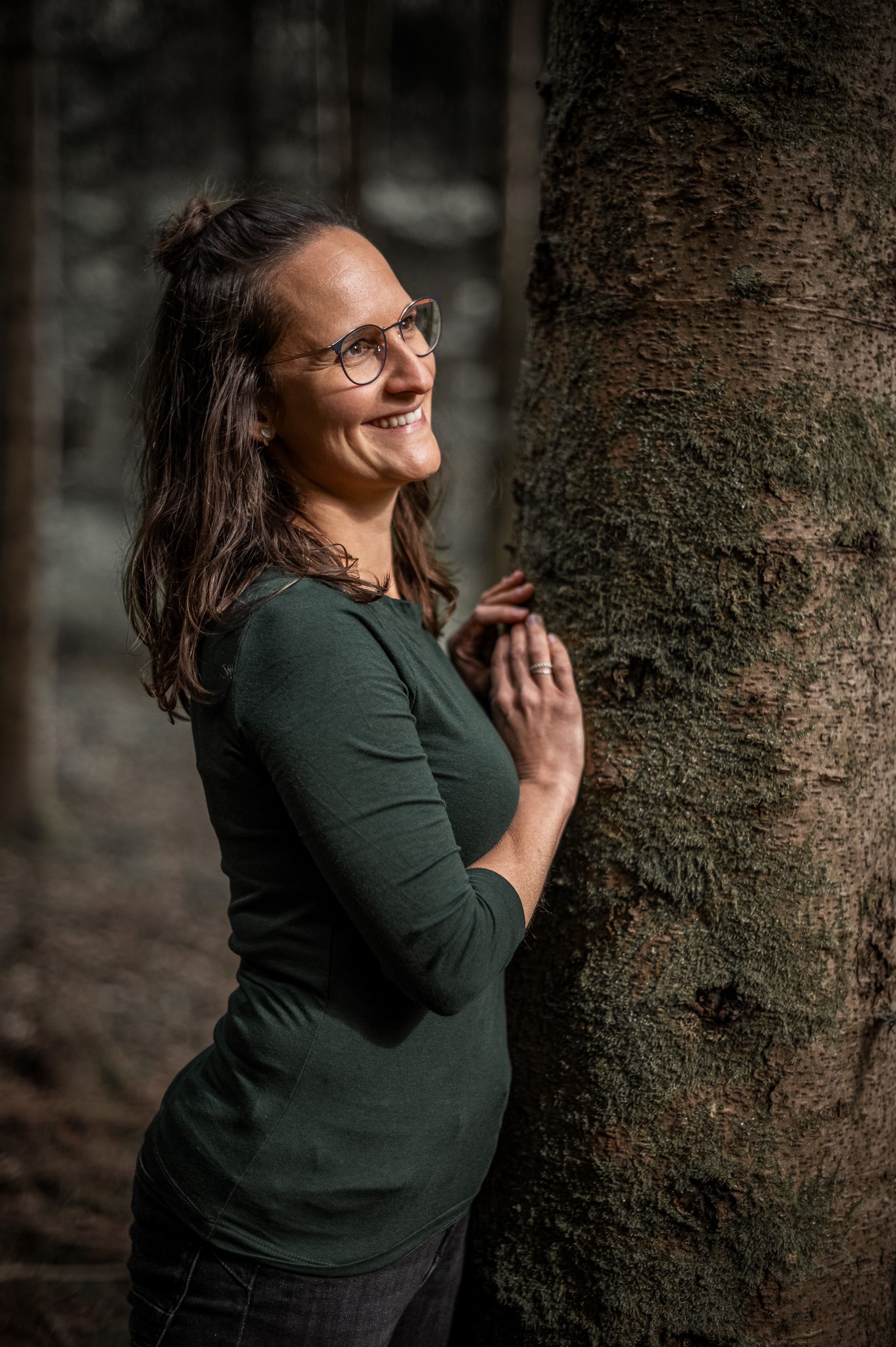 Sara lehnt zwischen zwei Baumstämmen im Wald und schaut lächelnd nach oben