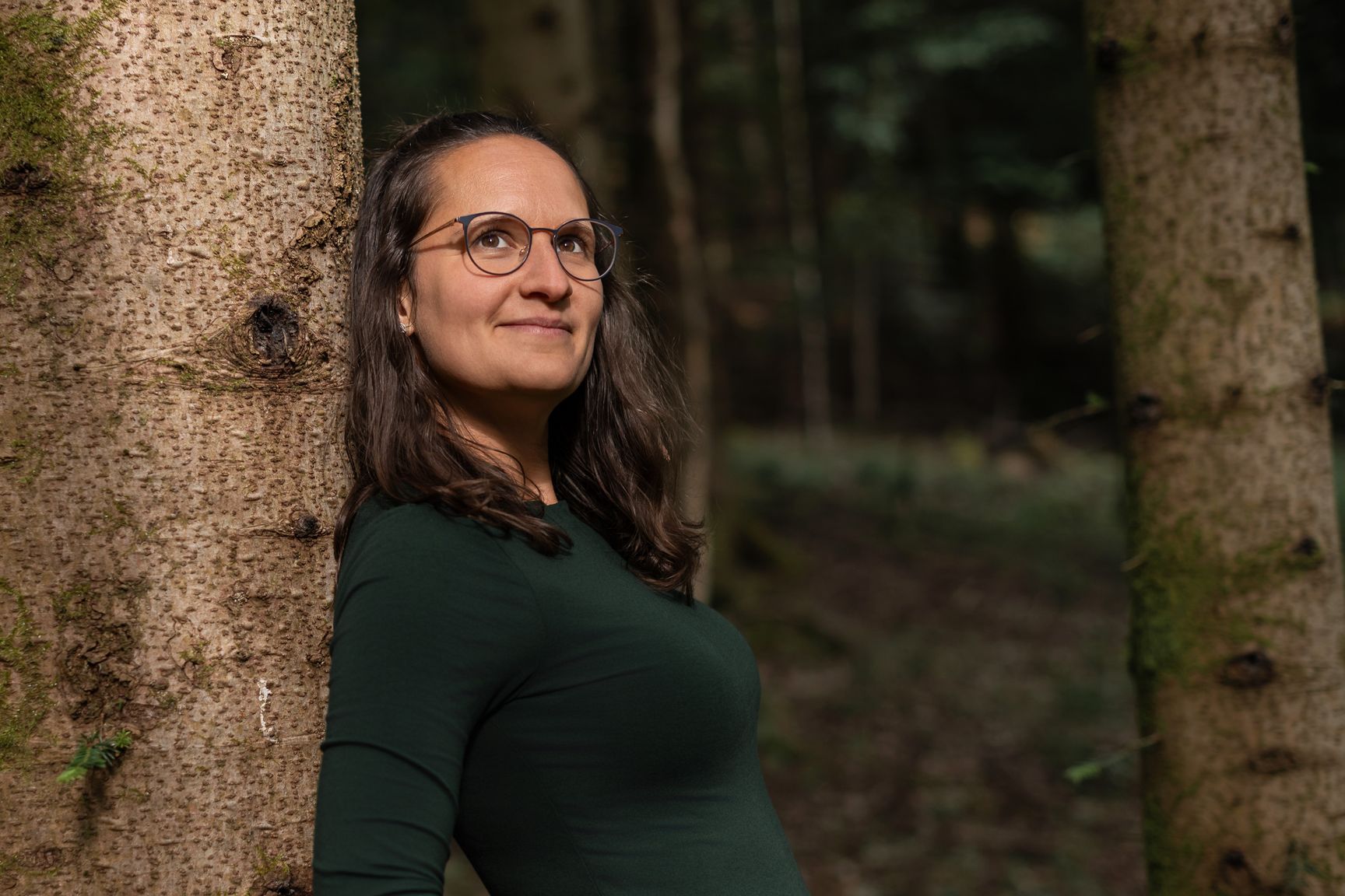 Sara lehnt zwischen zwei Baumstämmen im Wald und schaut lächelnd nach oben