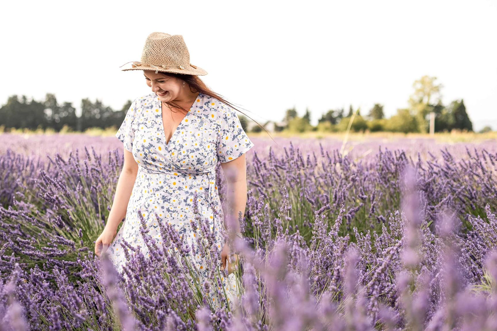 Frau im geblümten Sommerkleid mit Strohhut geht durch ein blühendes Lavendelfeld