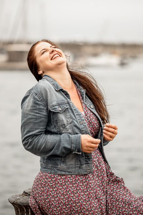 Frau in Jeansjacke lacht am Wasser, im Hintergrund ein Hafen
