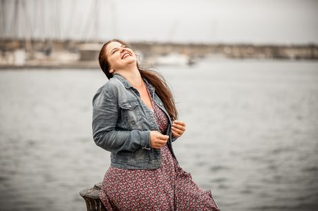 Frau in Jeansjacke lacht am Wasser, im Hintergrund ein Hafen