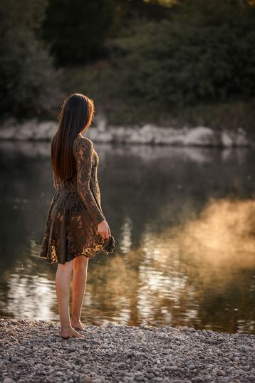 Frau sitzt im Profil am Kiesufer eines Flusses, die Abendsonne spiegelt sich im Wasser