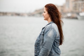 Frau in Jeansjacke steht im Profil am Ufer, der Wind bewegt ihr Haar