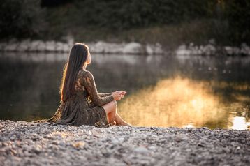 Frau sitzt im Profil am Kiesufer eines Flusses, die Abendsonne spiegelt sich im Wasser