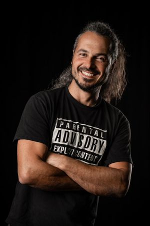 Mann im schwarzen T-Shirt steht mit verschränkten Armen vor dunklem Hintergrund