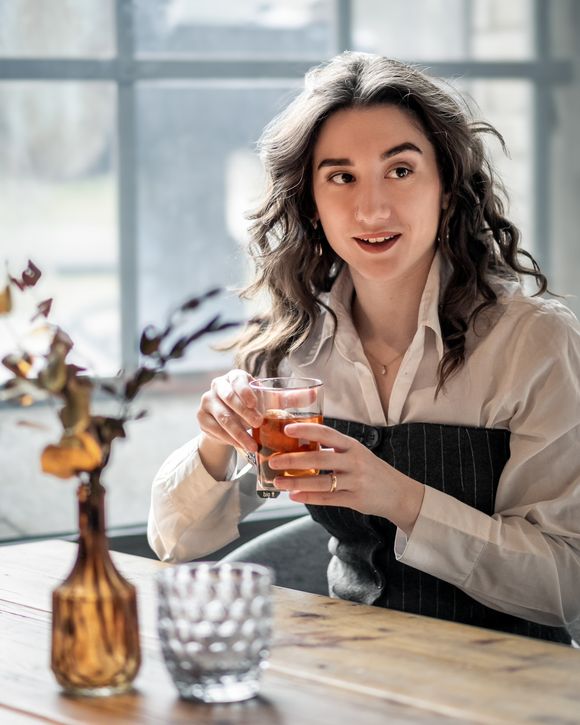 Frau mit einem Glas Tee sitzt an einem Holztisch am Fenster