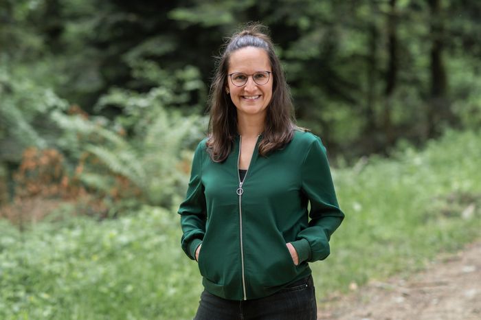 Sara mit Brille und grüner Jacke steht lächelnd auf einem Waldweg