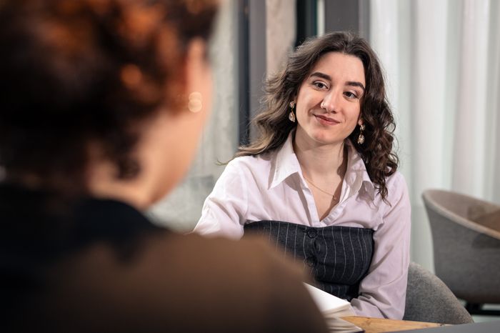 Simona in weisser Bluse sitzt am Tisch und hört ihrem Gegenüber lächelnd zu