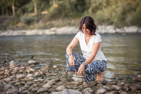 Frau kniet am Kiesufer eines Flusses und berührt mit der Hand das Wasser