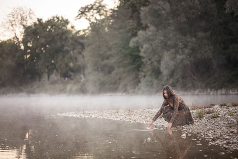 Frau kniet am Kiesufer eines Flusses und berührt mit der Hand das Wasser