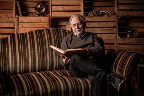 Älterer Mann sitzt mit einem Buch auf einem gestreiften Sofa in einer Scheune
