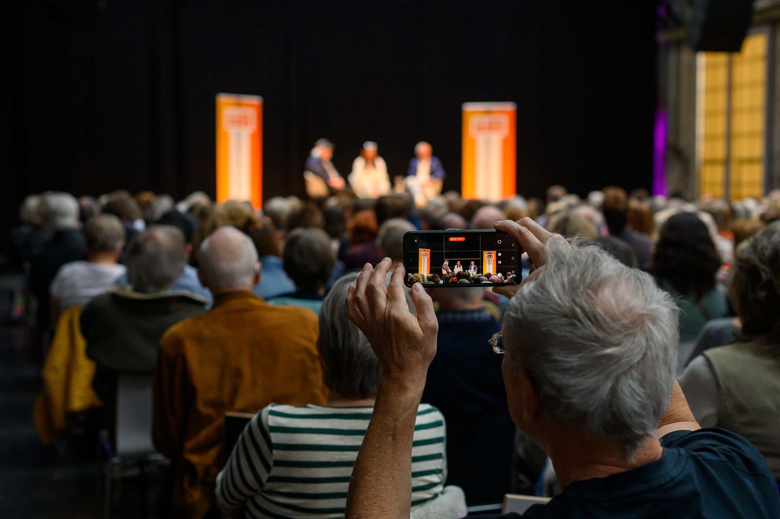Publikum von hinten, auf der Bühne ein Podium zwischen orangen Stelen