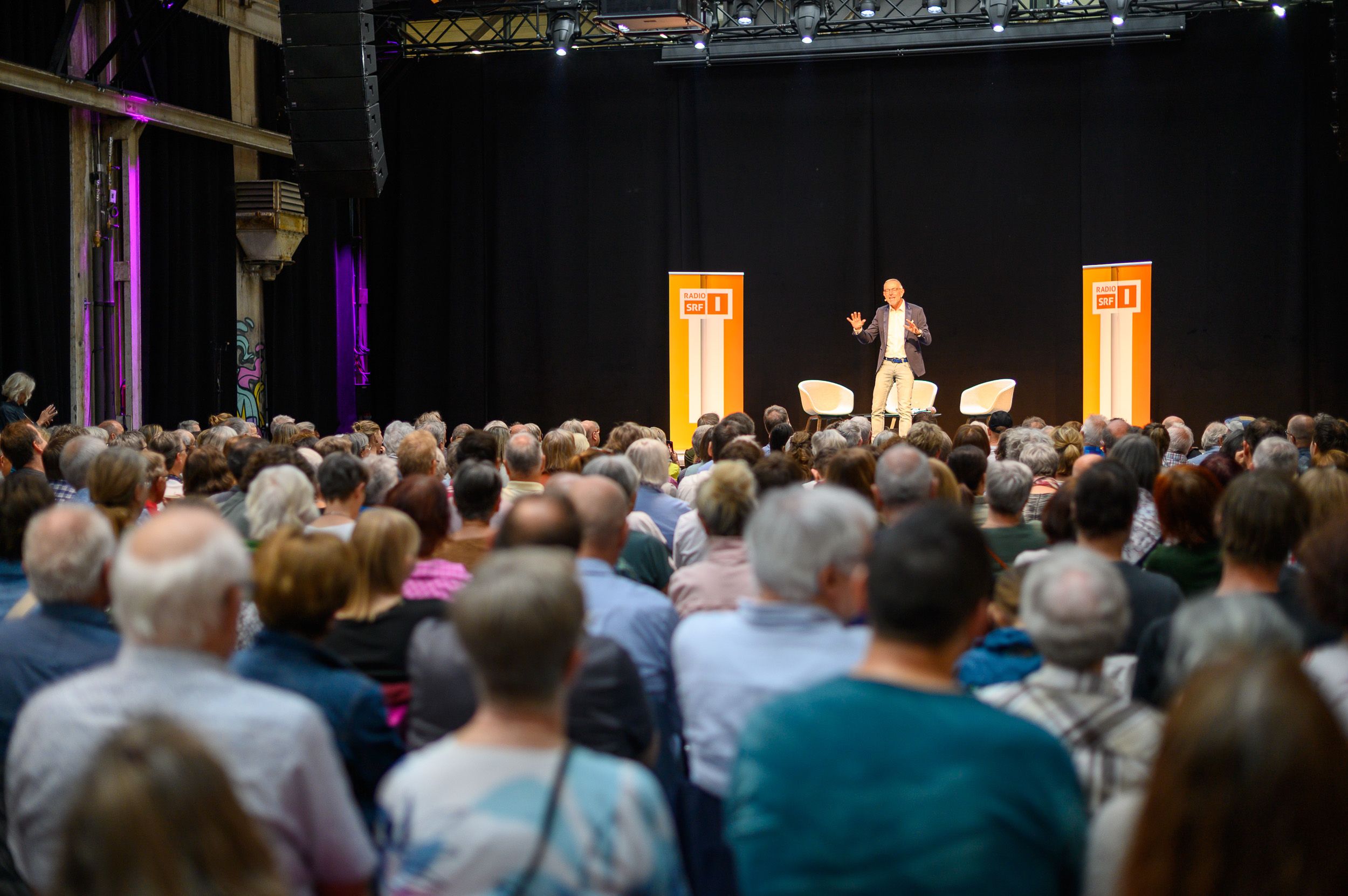 Volles Publikum von hinten, auf der Bühne eine Rednerin zwischen orangen Stelen