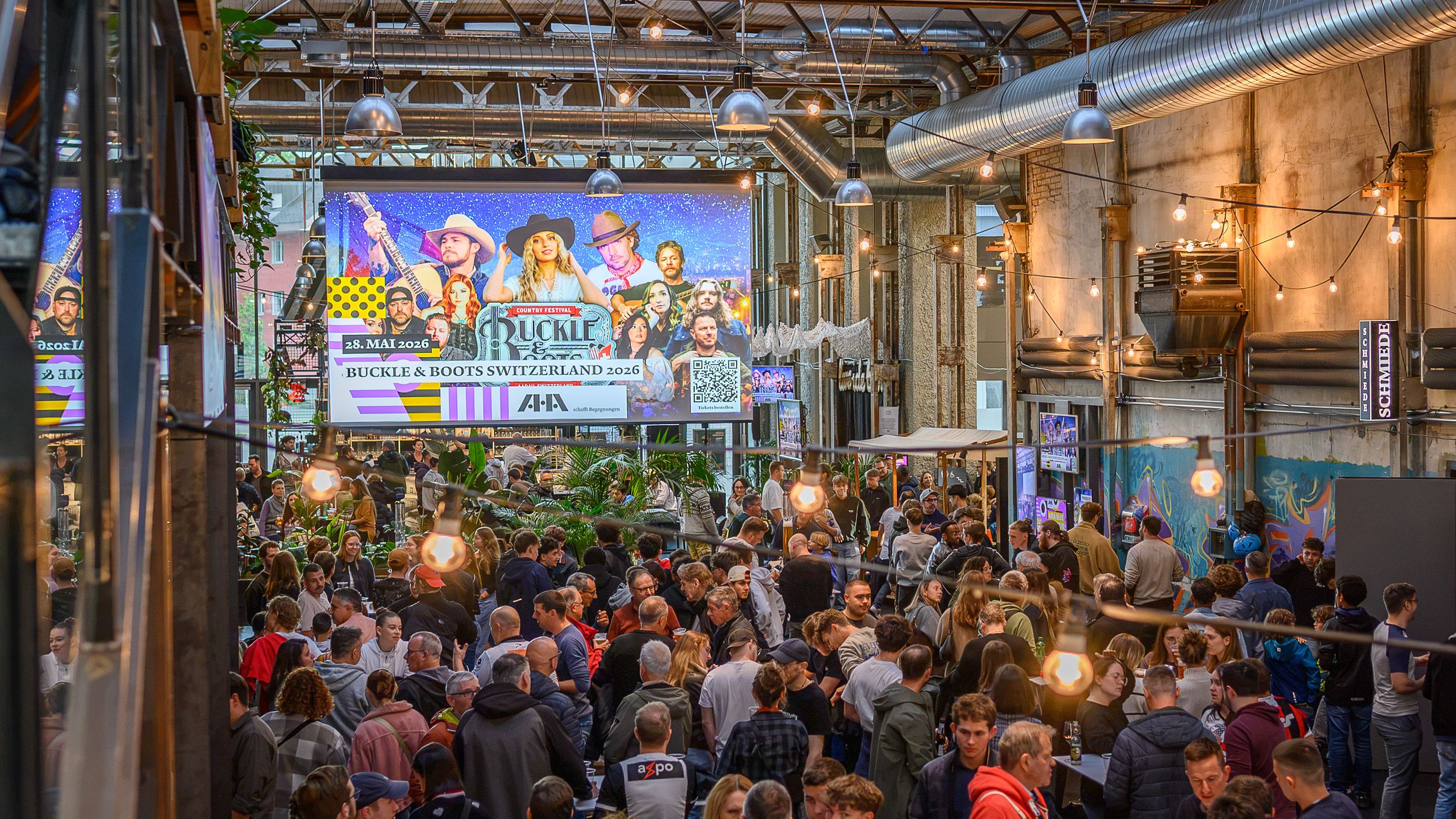 Volle Industriehalle mit grosser Bühne und Leinwand, dichtes Publikum davor