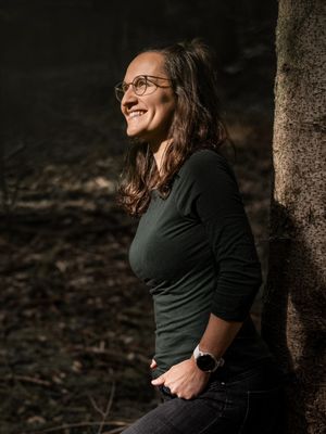 Frau mit Brille steht neben einem Baum im Wald und lächelt