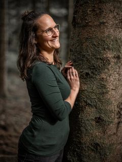 Frau mit Brille lehnt an einem Baumstamm, die Hände vor der Brust