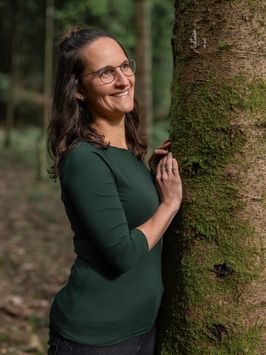 Sara lehnt mit beiden Händen an einem bemoosten Baumstamm und lächelt