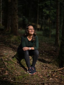 Sara sitzt auf einem Baumstrunk im Wald, die Hände auf den Knien