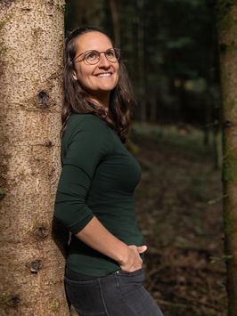 Sara lehnt an einem Baumstamm, die Hände in den Hosentaschen, und schaut nach oben