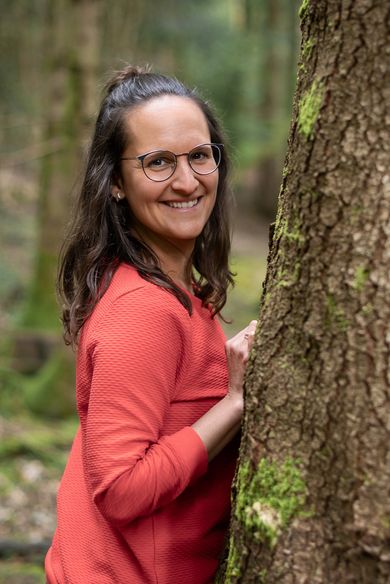 Sara steht an einem bemoosten Baumstamm und lächelt in die Kamera