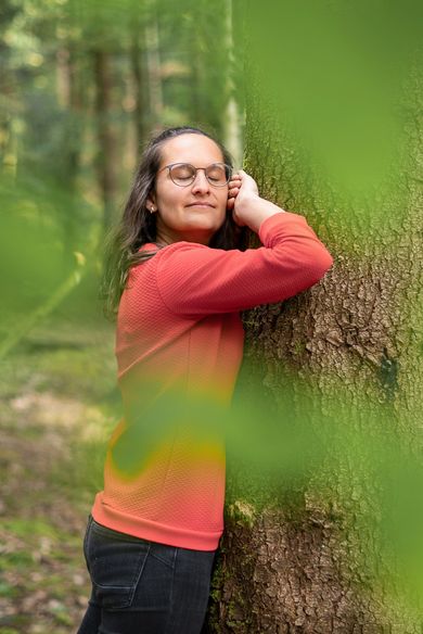 Sara lehnt den Kopf an einen bemoosten Stamm, die Augen geschlossen, im Vordergrund unscharfes Grün