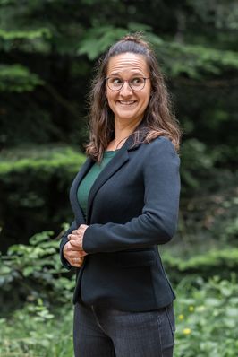 Sara im dunklen Blazer steht im Grünen, die Hände vor dem Körper