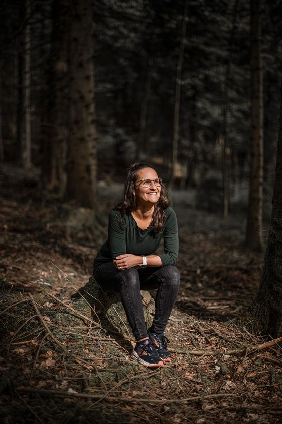 Sara sitzt auf einem Baumstrunk im Wald und schaut lächelnd nach oben