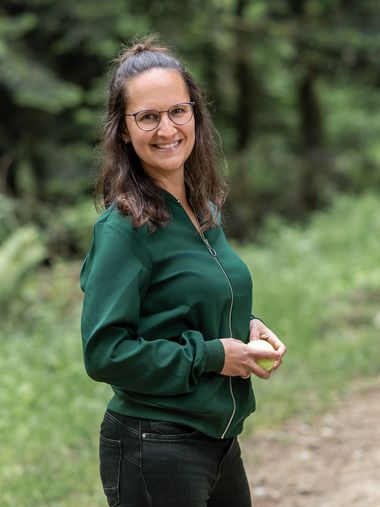 Sara steht auf einem Waldweg, einen Tennisball in der Hand, und lächelt in die Kamera