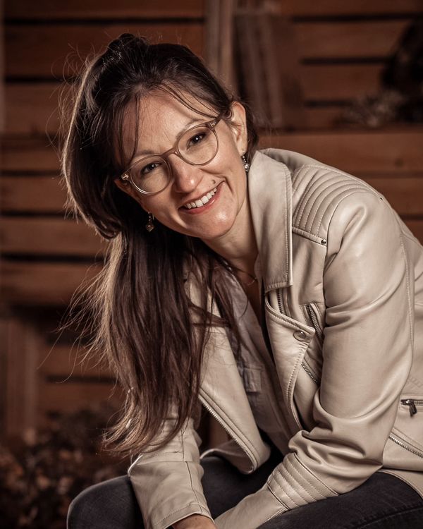 Seraina Wild mit Brille in heller Lederjacke, kniend vor einer Holzwand