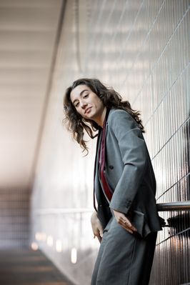 Simona im grauen Anzug lehnt an einer hellen Wand