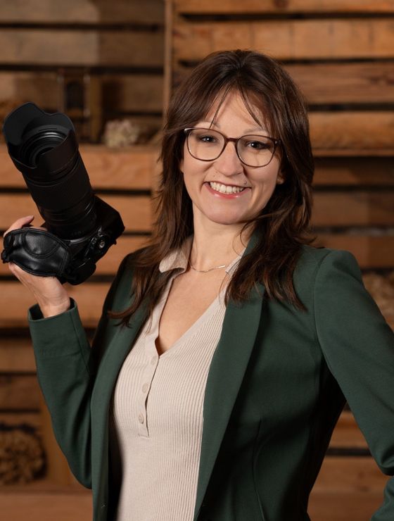 Seraina Wild mit Brille im dunkelgrünen Blazer hält lachend ihre Kamera hoch, Holzkisten im Hintergrund